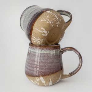 May include: Two handmade ceramic mugs stacked on top of each other. The mugs have a two-tone design with a brown base and a purple and white upper section. White floral patterns decorate the brown sections. Each mug has a rounded handle.