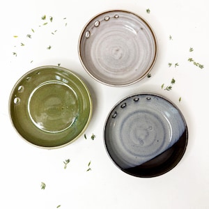 May include: Three circular ceramic bowls with a glossy finish. One bowl is olive green, one is light beige, and one is a gradient of blue and dark brown. Each bowl features a series of small, evenly spaced circular holes along one side of the rim.