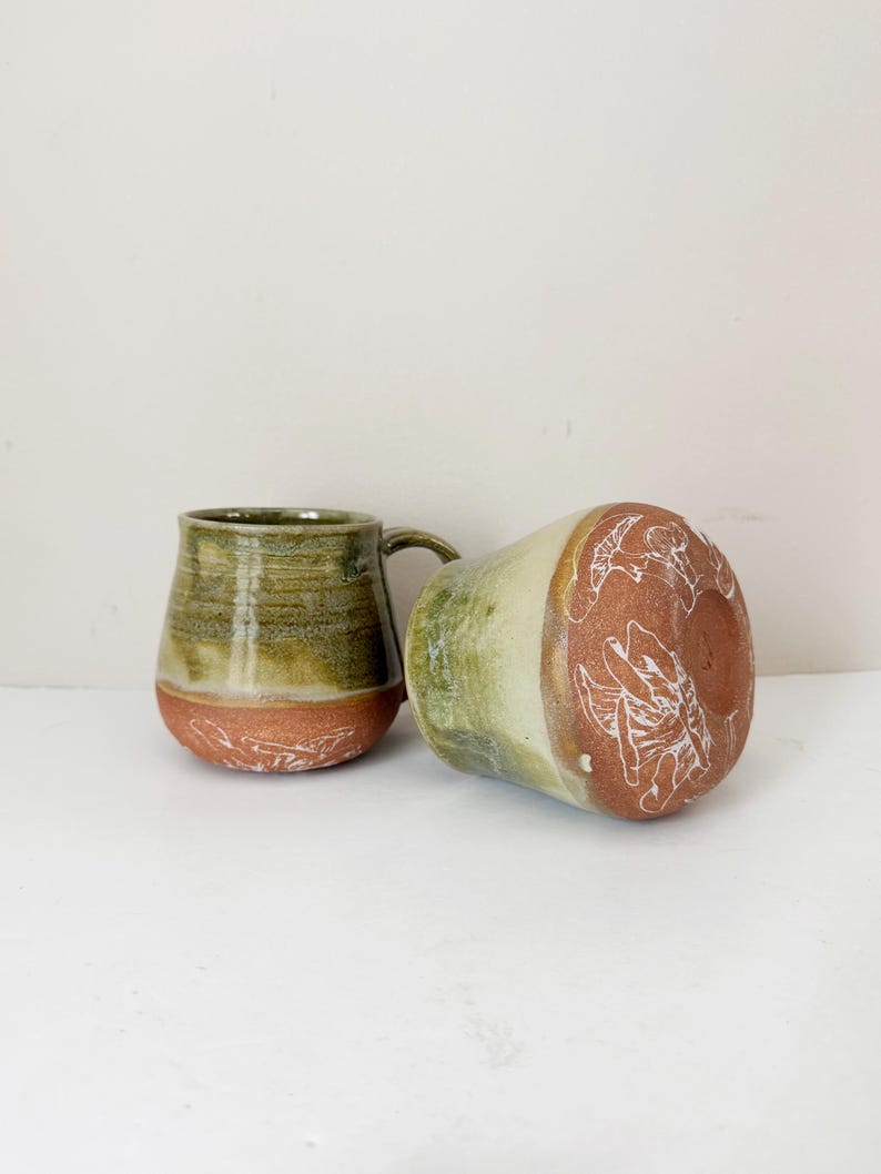 May include: Two handmade ceramic mugs. One is upright, the other is on its side. The mugs have a green and brown glaze with white etched designs on the base. The mugs are a unique addition to any kitchen.