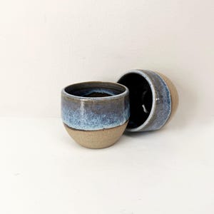 May include: Two small ceramic cups with a rounded shape. The lower portion of each cup features a natural, unglazed tan clay finish, while the upper portion is coated in a glossy, reactive glaze that transitions from deep navy blue to light blue.