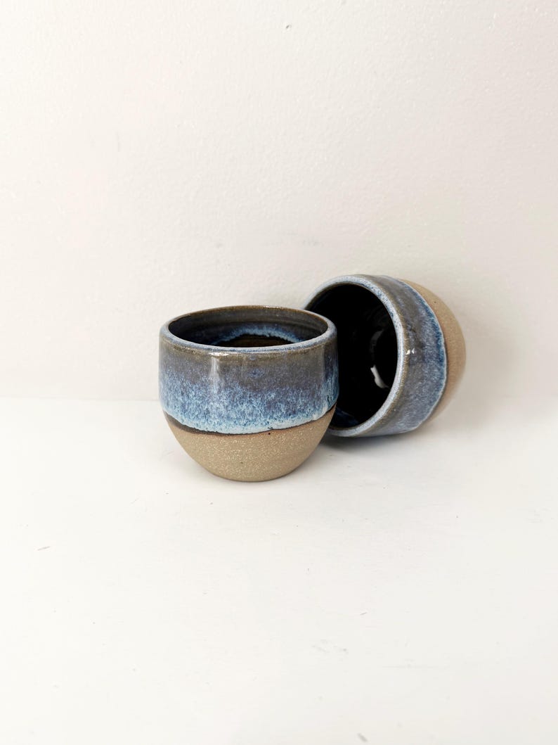 May include: Two small ceramic cups with a rounded shape. The lower portion of each cup features a natural, unglazed tan clay finish, while the upper portion is coated in a glossy, reactive glaze that transitions from deep navy blue to light blue.