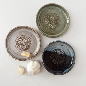 May include: Three circular ceramic grater plates with raised central textures for grating garlic. One plate is sage green, one is light gray, and one is dark blue and black. Two garlic cloves and one whole garlic bulb are placed nearby.