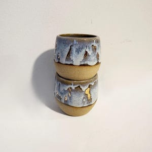May include: Two stacked ceramic cups with a rounded shape. The bottom halves are unglazed, textured tan clay, while the top halves feature a glossy, drippy blue and white glaze with small metallic gold accents visible on the surface of the lower cup.