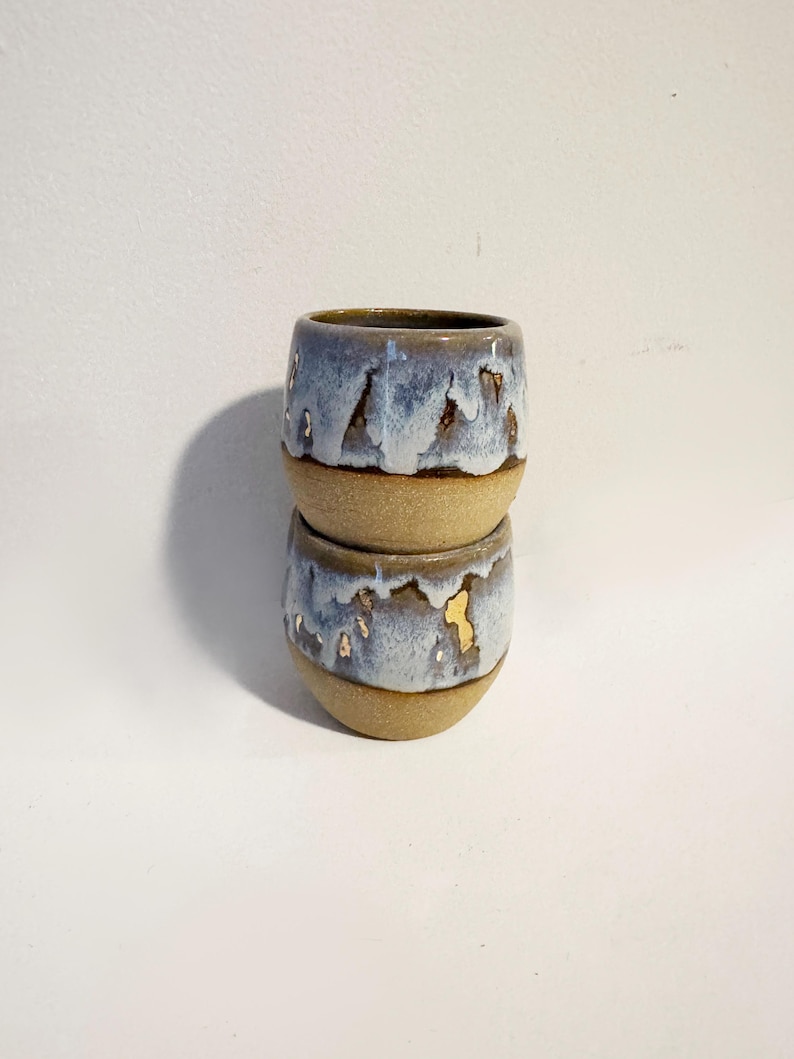 May include: Two stacked ceramic cups with a rounded shape. The bottom halves are unglazed, textured tan clay, while the top halves feature a glossy, drippy blue and white glaze with small metallic gold accents visible on the surface of the lower cup.