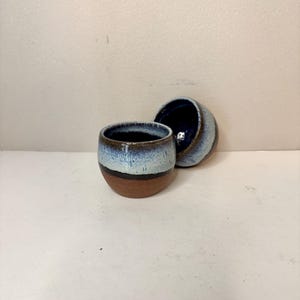 May include: Two small, round ceramic cups featuring a reactive glaze. The lower portion of each cup is unglazed, showing a natural brown clay texture, while the upper section transitions from a dark band into a speckled light blue and white finish.