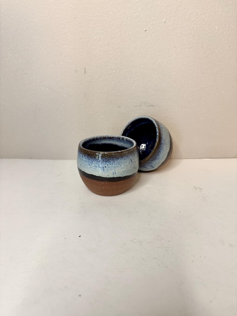 May include: Two small, round ceramic cups featuring a reactive glaze. The lower portion of each cup is unglazed, showing a natural brown clay texture, while the upper section transitions from a dark band into a speckled light blue and white finish.
