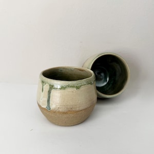 May include: Two ceramic cups with a rounded shape. The exterior features a smooth, cream-coloured glaze on the upper half with green drips, while the lower half is a textured, unglazed brown clay. The interior is finished in a dark green glaze.
