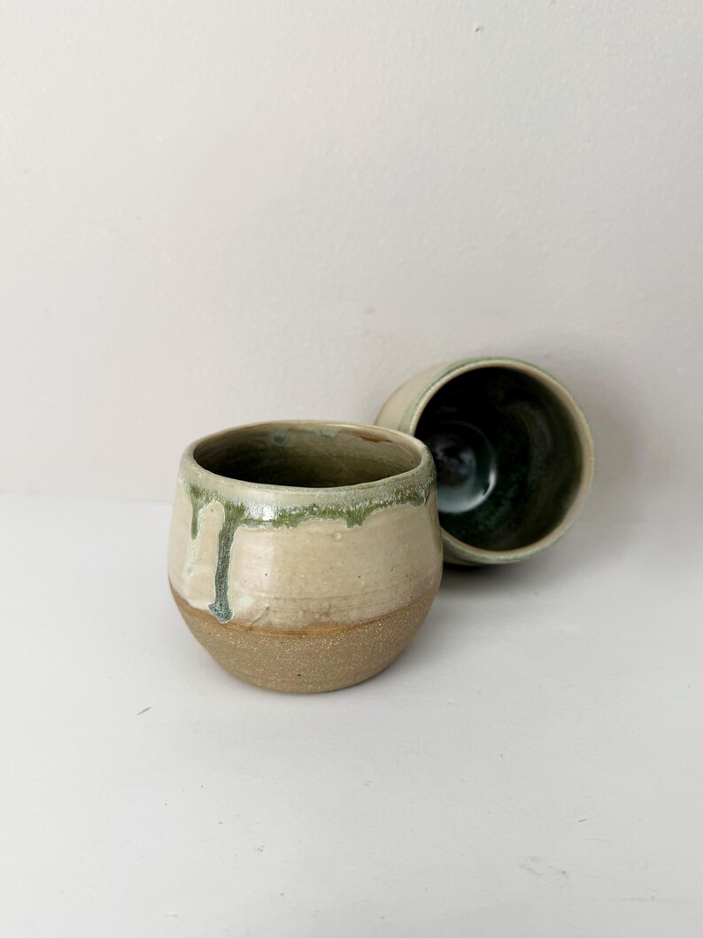 May include: Two ceramic cups with a rounded shape. The exterior features a smooth, cream-coloured glaze on the upper half with green drips, while the lower half is a textured, unglazed brown clay. The interior is finished in a dark green glaze.