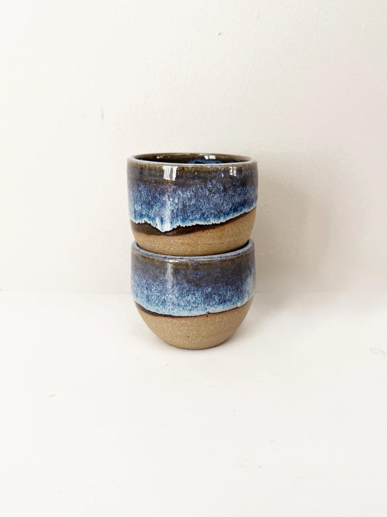 May include: Two stacked ceramic cups featuring a smooth, unglazed tan base and a reactive glaze top in shades of deep navy blue and lighter sky blue. The glaze drips down the sides, creating a textured transition between the blue and tan sections.