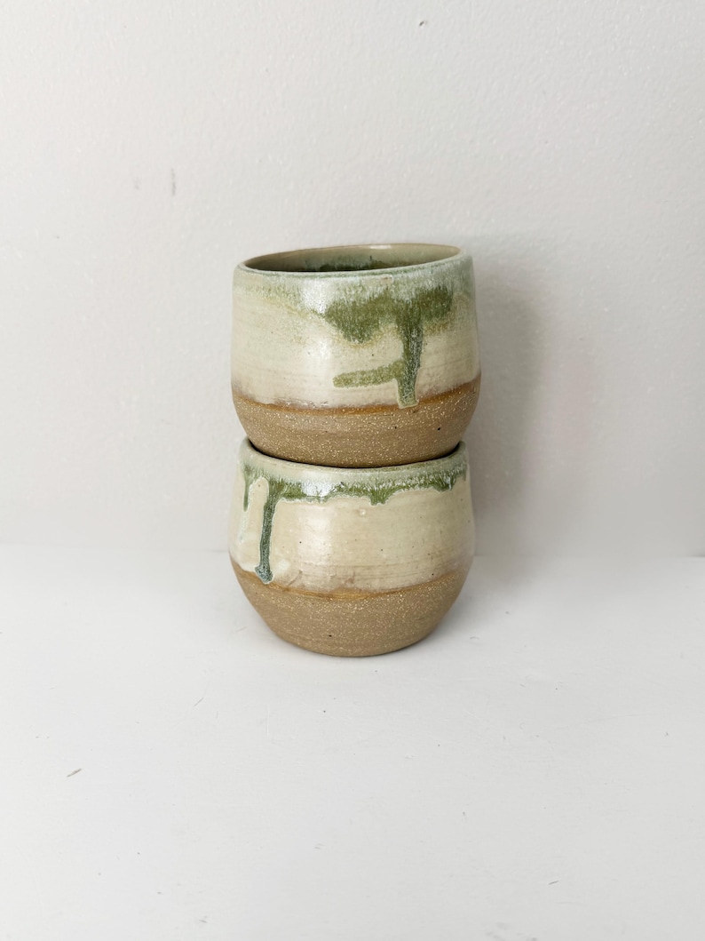 May include: Two handmade ceramic cups stacked on top of each other. The cups have a beige base with a green and white glaze that drips down the sides. The cups are likely used for drinking beverages.