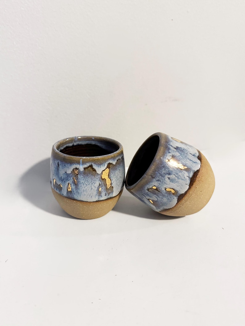 May include: Two small ceramic cups with a rounded shape. The lower portion features a textured, matte beige finish, while the upper section is glazed in a mottled blue and white pattern with small, irregular gold-coloured accents throughout the glaze.