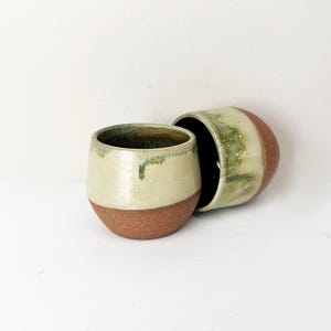 May include: Two rounded ceramic cups featuring a textured, unglazed terracotta base and a smooth, glossy cream-coloured upper section with green glaze drips running down the sides. The interior of the cups is finished in a dark, glossy glaze.