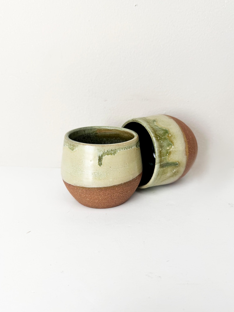 May include: Two rounded ceramic cups featuring a textured, unglazed terracotta base and a smooth, glossy cream-coloured upper section with green glaze drips running down the sides. The interior of the cups is finished in a dark, glossy glaze.
