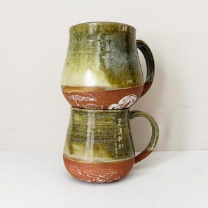 May include: Two handmade ceramic mugs stacked on top of each other. The mugs have a green and brown glaze with white mushroom designs around the base. Each mug has a dark brown handle.