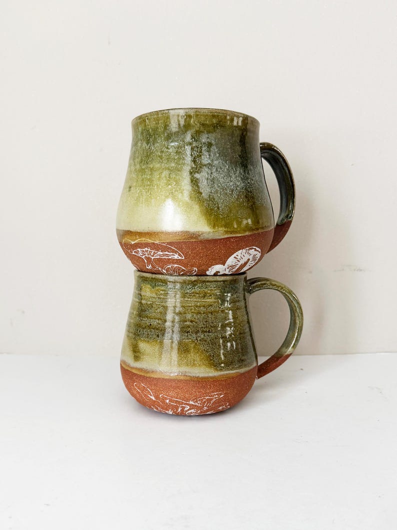May include: Two handmade ceramic mugs stacked on top of each other. The mugs have a green and brown glaze with white mushroom designs around the base. Each mug has a dark brown handle.