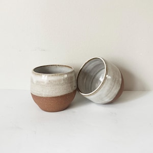 May include: Two small, rounded ceramic cups featuring a two-tone design. The lower portion of each cup is made of unglazed, speckled terracotta-coloured clay, while the upper portion is finished with a smooth, glossy, off-white glaze.