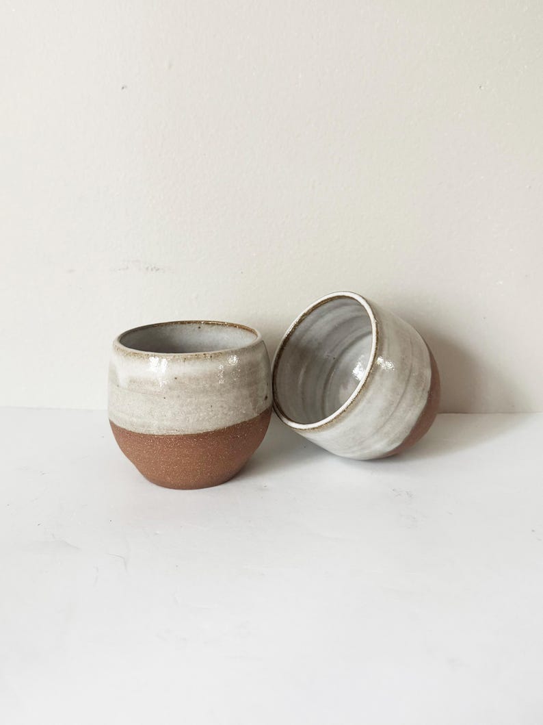 May include: Two small, rounded ceramic cups featuring a two-tone design. The lower portion of each cup is made of unglazed, speckled terracotta-coloured clay, while the upper portion is finished with a smooth, glossy, off-white glaze.