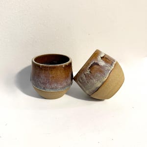 May include: Two ceramic cups with a rounded shape. The upper portions feature a reactive glaze in shades of brown, tan, and hints of purple, while the lower portions remain an unglazed, textured beige clay. One cup stands upright, and the other rests on its side.