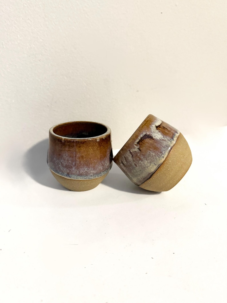 May include: Two ceramic cups with a rounded shape. The upper portions feature a reactive glaze in shades of brown, tan, and hints of purple, while the lower portions remain an unglazed, textured beige clay. One cup stands upright, and the other rests on its side.