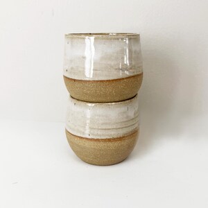 May include: Two stacked ceramic cups featuring a two-tone design. The upper portion of each cup is finished with a smooth, glossy off-white glaze, while the lower portion displays a textured, speckled tan clay surface with a visible horizontal line.