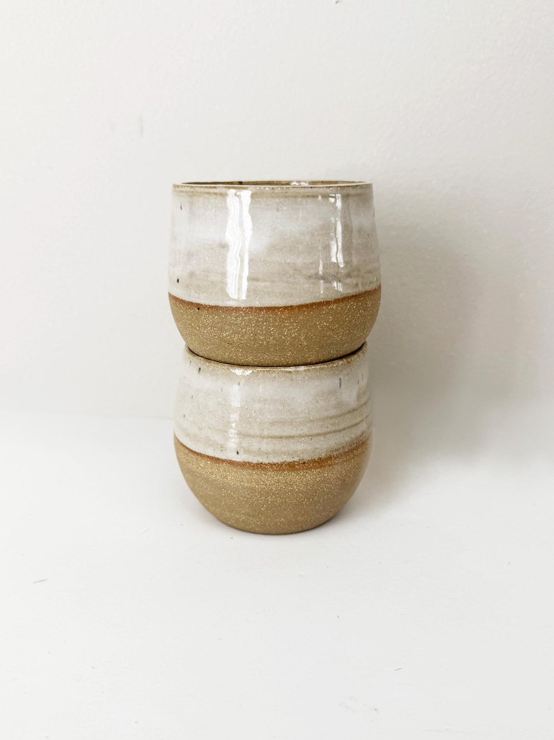 May include: Two stacked ceramic cups featuring a two-tone design. The upper portion of each cup is finished with a smooth, glossy off-white glaze, while the lower portion displays a textured, speckled tan clay surface with a visible horizontal line.