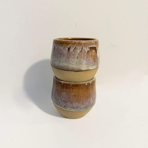 May include: Two stacked ceramic cups featuring a rounded shape and a two-tone finish. The upper portion of each cup has a glossy, mottled glaze in shades of brown, mauve, and cream, while the lower portion displays a textured, matte tan finish.