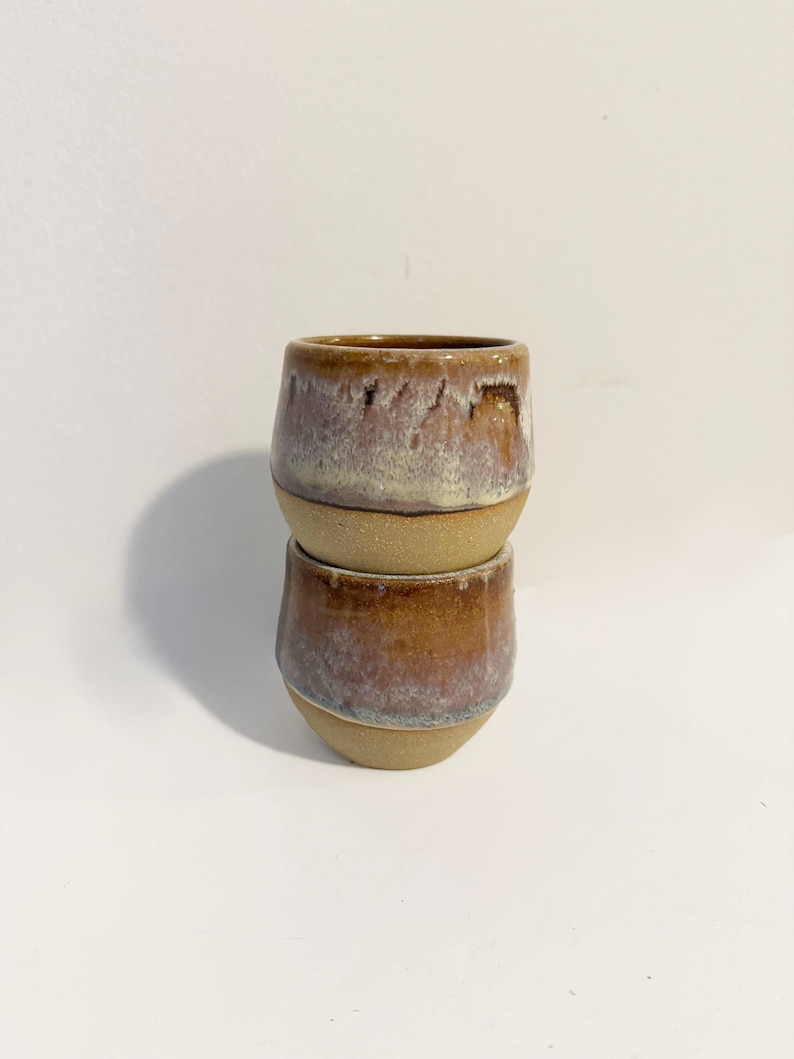 May include: Two stacked ceramic cups featuring a rounded shape and a two-tone finish. The upper portion of each cup has a glossy, mottled glaze in shades of brown, mauve, and cream, while the lower portion displays a textured, matte tan finish.