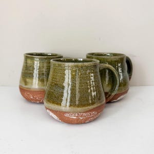 May include: Three handmade ceramic mugs. The mugs have a rounded shape with a dark green glaze on the upper portion and a reddish-brown base. The mugs have a handle and are suitable for coffee or tea.
