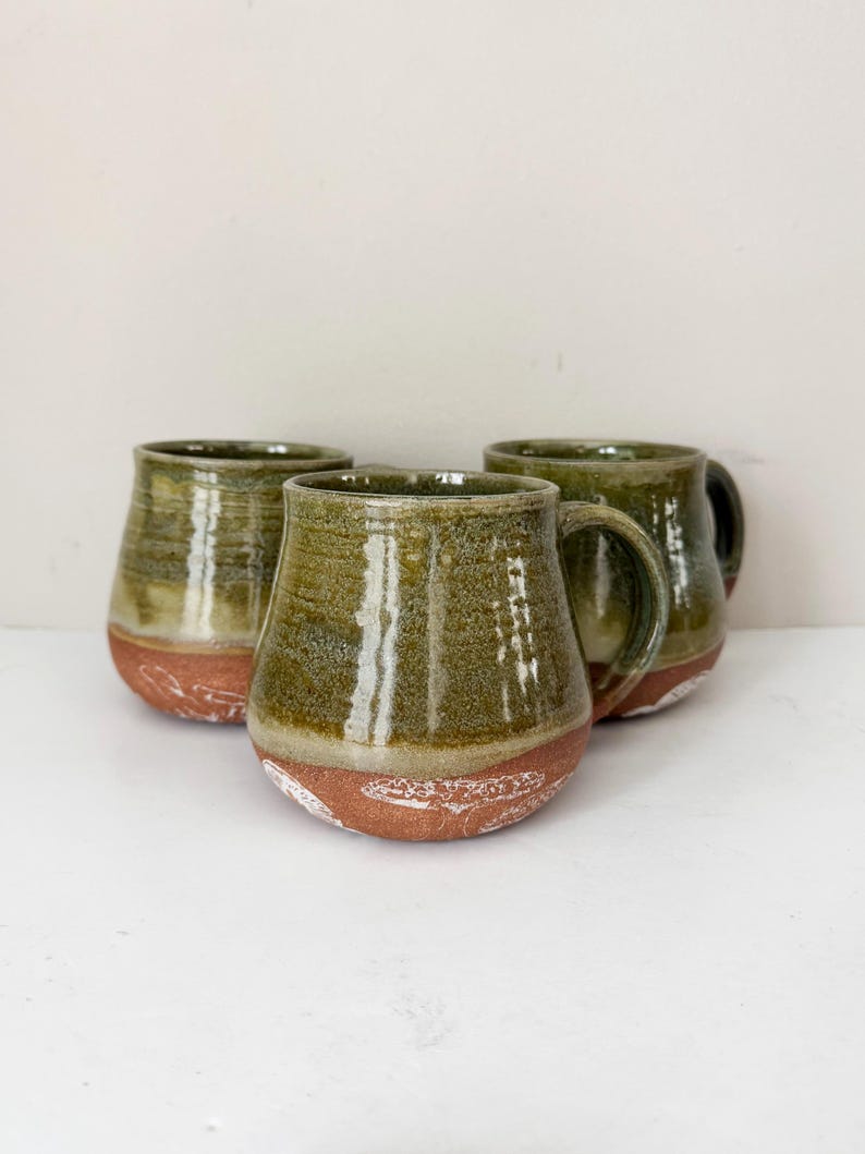 May include: Three handmade ceramic mugs. The mugs have a rounded shape with a dark green glaze on the upper portion and a reddish-brown base. The mugs have a handle and are suitable for coffee or tea.