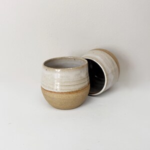 May include: Two ceramic tumblers with a rounded shape. The upper portion of each tumbler features a smooth, off-white glaze, while the lower portion displays a textured, speckled tan clay finish. The interior of the tumblers is dark brown.