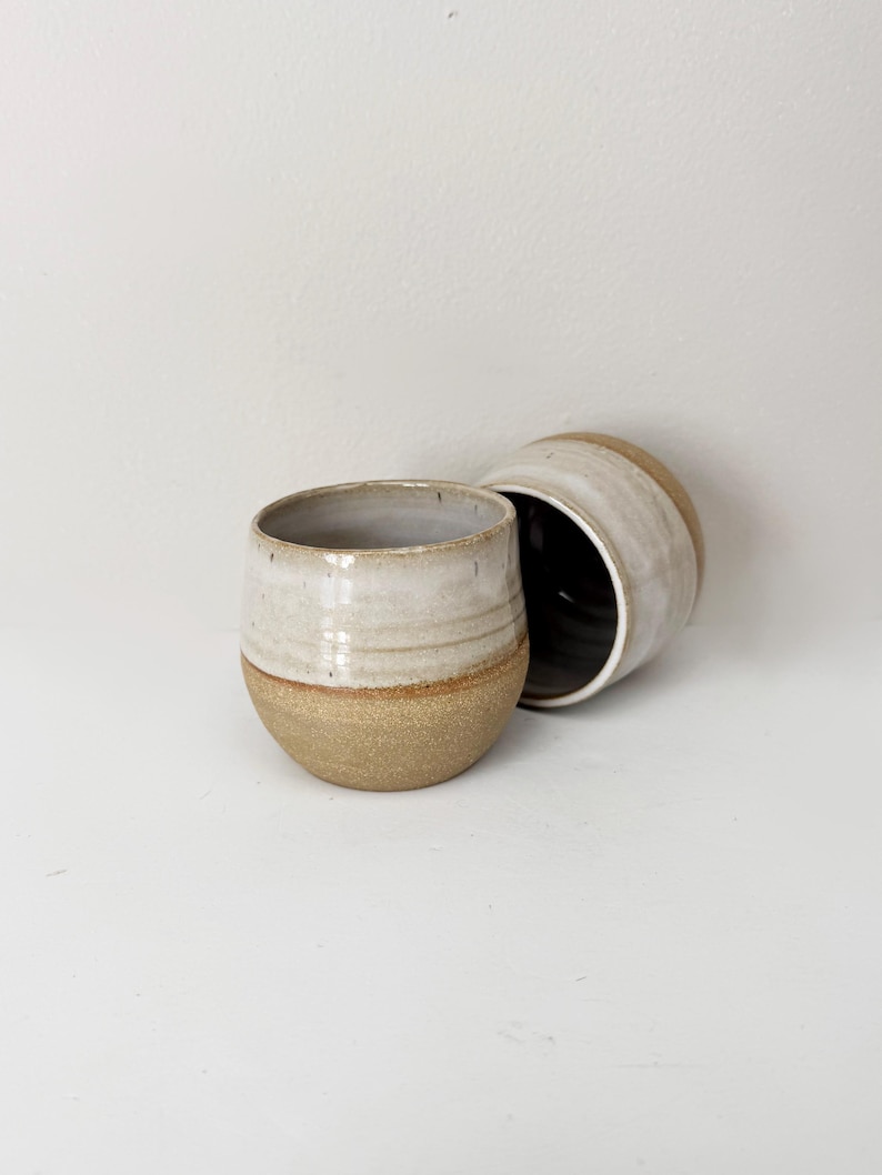 May include: Two ceramic tumblers with a rounded shape. The upper portion of each tumbler features a smooth, off-white glaze, while the lower portion displays a textured, speckled tan clay finish. The interior of the tumblers is dark brown.