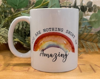 May include: White ceramic mug with a curved handle. The mug features a watercolor rainbow design with the words "YOU ARE NOTHING SHORT OF Amazing" in black script. The mug is on a wooden tray.
