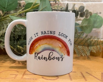 May include: White ceramic mug with a curved handle. The mug features a watercolor rainbow design with the words "When it rains look for Rainbows" in black script. The mug is set against a background of greenery.
