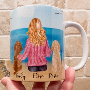May include: White ceramic mug featuring a beach scene with a woman and two dogs. The woman has blonde hair and wears a pink sweater. The dogs are brown and tan. The names "Ruby", "Elise", and "Rosie" are written on the mug.