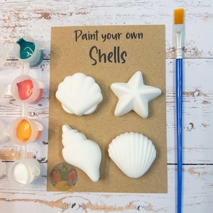 May include: A craft kit featuring the text "Paint your own Shells" includes four white seashell shapes, paint pots with various colours, and a paintbrush with a blue handle. The kit is designed for painting and decorating.