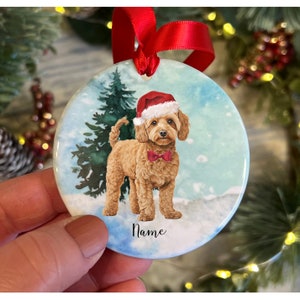 May include: A circular Christmas ornament depicting a light brown dog in a Santa hat and red bow tie, set against a winter scene with a Christmas tree. The word "Name" is at the bottom. A red ribbon is attached for hanging.