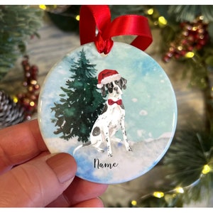 May include: Circular Christmas ornament depicting a dog in a Santa hat and bow tie, set in a snowy scene with a fir tree. The ornament has a red ribbon for hanging and the word "Name" is written on the bottom.