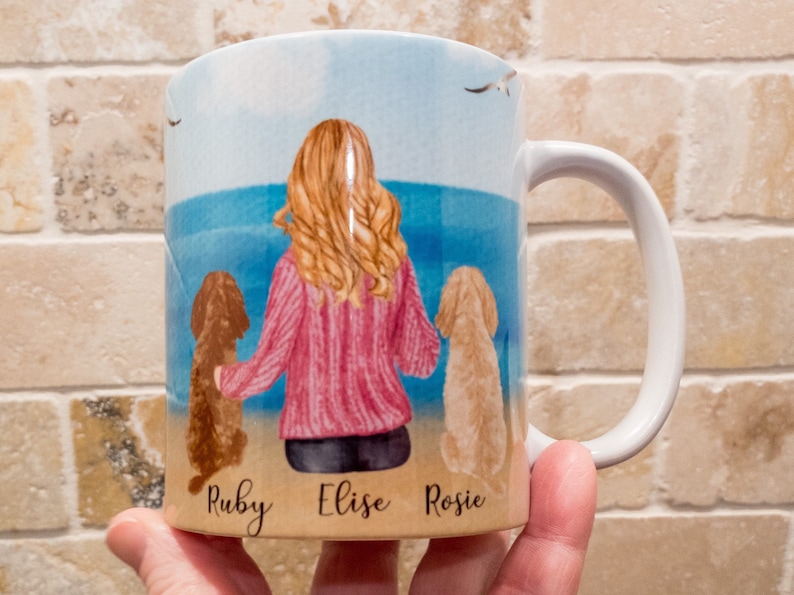 May include: White ceramic mug featuring a beach scene with a woman and two dogs. The woman has blonde hair and wears a pink sweater. The dogs are brown and tan. The names "Ruby", "Elise", and "Rosie" are written on the mug.