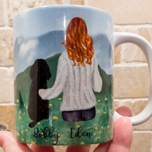 May include: White ceramic mug featuring a watercolor illustration of a person with red hair and a black dog in a field. The names "Bobby" and "Eden" are written at the bottom. The mug has a white handle and a blue sky background.