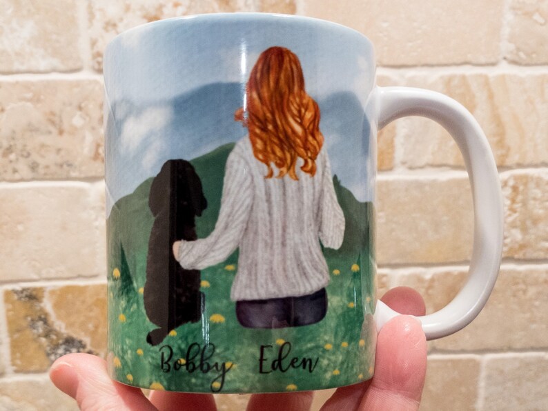 May include: White ceramic mug featuring a watercolor illustration of a person with red hair and a black dog in a field. The names "Bobby" and "Eden" are written at the bottom. The mug has a white handle and a blue sky background.