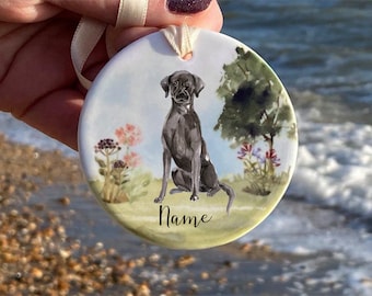 May include: Round ceramic ornament featuring a watercolor painting of a black dog sitting in a field with flowers and a tree. The word "Name" is written in script below the dog. A cream-colored ribbon is attached for hanging.