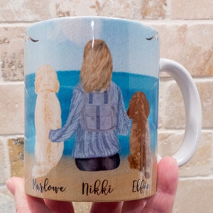 May include: White ceramic mug featuring a watercolor illustration of a person with two dogs on a beach. The names "Marlowe", "Nikki", and "Elfie" are printed below the dogs. The background shows a blue sky and ocean.