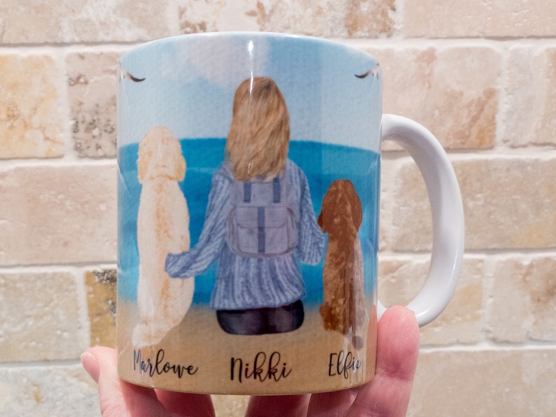 May include: White ceramic mug featuring a watercolor illustration of a person with two dogs on a beach. The names "Marlowe", "Nikki", and "Elfie" are printed below the dogs. The background shows a blue sky and ocean.