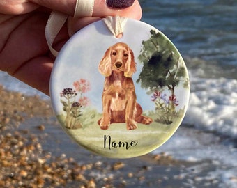 May include: Round ceramic ornament featuring a watercolor painting of a Cocker Spaniel dog sitting in a field with flowers and a tree. The dog is light brown with long ears. The word "Name" is written in script at the bottom. A cream ribbon is attached for hanging.