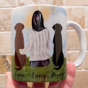 May include: White ceramic mug featuring a watercolor illustration of a person with two dogs. The dogs are a brown Labrador and a black Labrador. The names "Coco", "Lucy", and "Poppy" are written below the dogs.