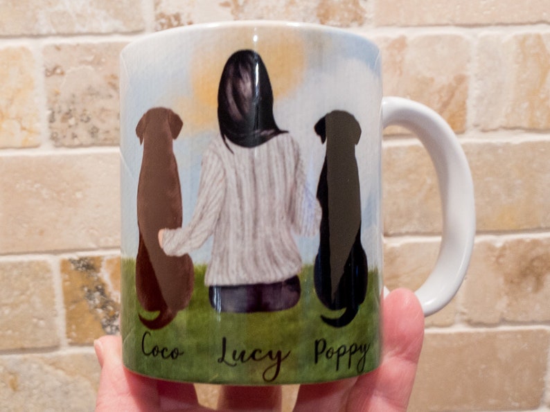 May include: White ceramic mug featuring a watercolor illustration of a person with two dogs. The dogs are a brown Labrador and a black Labrador. The names "Coco", "Lucy", and "Poppy" are written below the dogs.