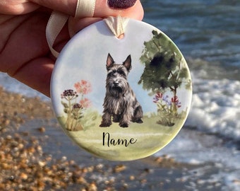 May include: A round ceramic ornament featuring a seated gray dog with a black face, ears, and nose. The dog is set against a backdrop of a grassy field, flowers, and a tree. The word "Name" is written below the dog.