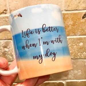 May include: White ceramic mug with a beach-themed design. The mug features a blue and orange watercolor background with the text "Life is better when I'm with my dog" in black script. A small dog graphic is also visible.