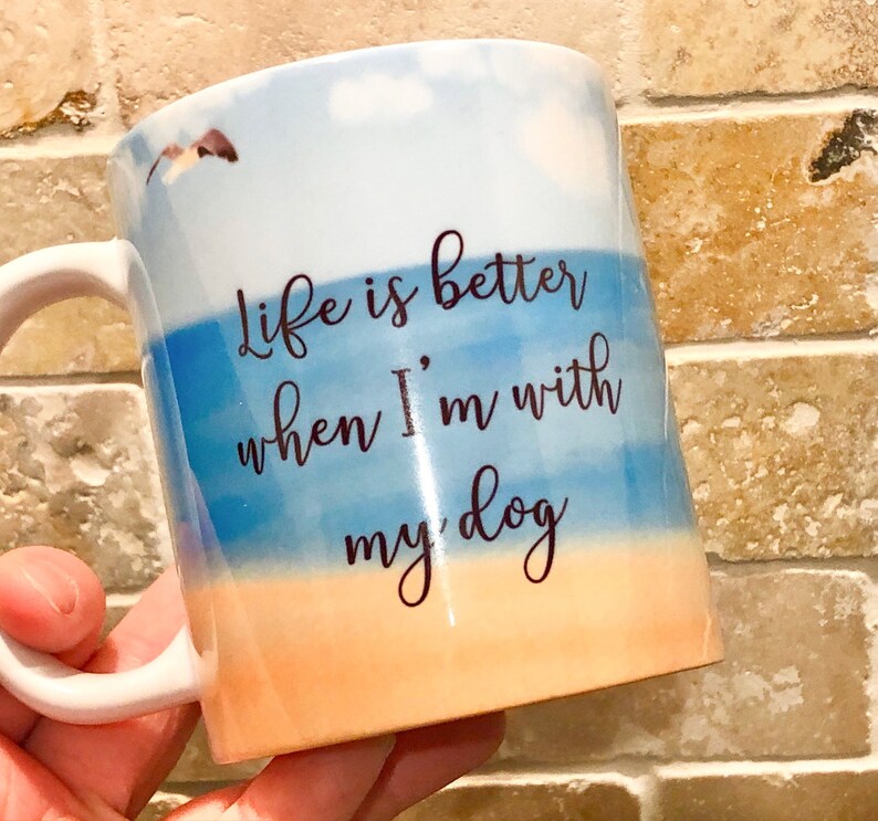May include: White ceramic mug with a beach-themed design. The mug features a blue and orange watercolor background with the text "Life is better when I'm with my dog" in black script. A small dog graphic is also visible.