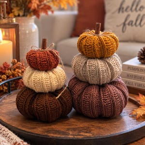 May include: Two stacks of crocheted pumpkins in varying sizes and colors, including shades of dark brown, cream, rust orange, mustard yellow, and muted mauve. Each pumpkin features a ribbed texture and a cinnamon stick stem tied with twine.